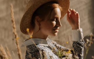 A woman sitting in a field with eyes closed touching her hat